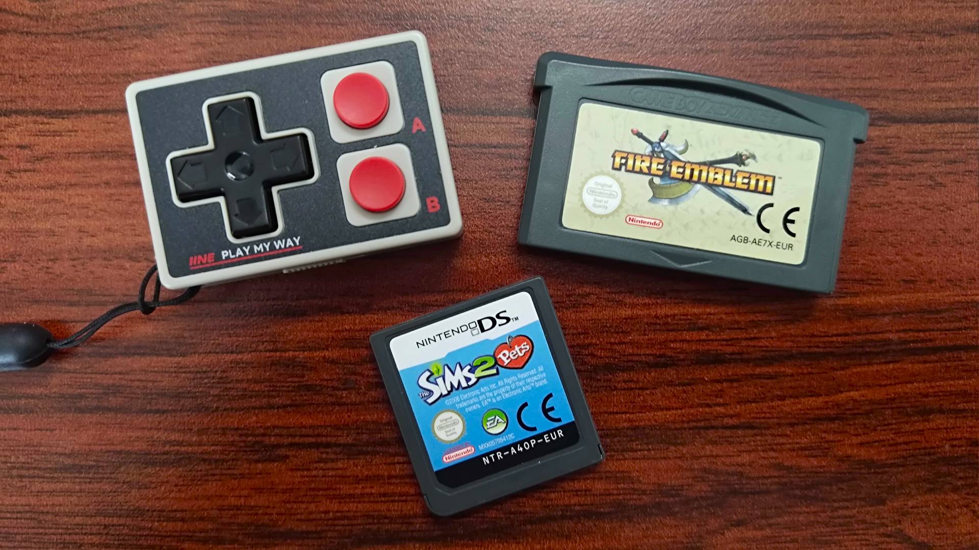 IINE Gamebrick Mini NES controller next to Fire Emblem Game Boy Advance cartridge and Sims 2 Pets for Nintendo DS on woodgrain desk.