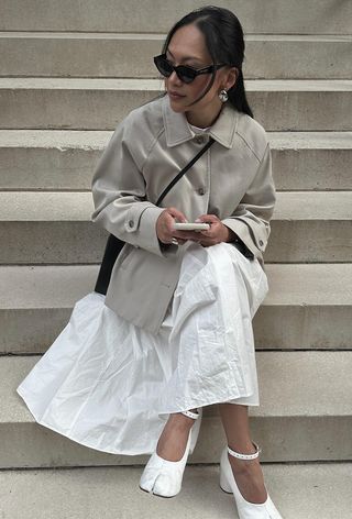 best cropped trench coat for women shown on a woman sitting outside on steps wearing a short tan trench over a white dress with white tabi toe pumps and a black crossbody bag