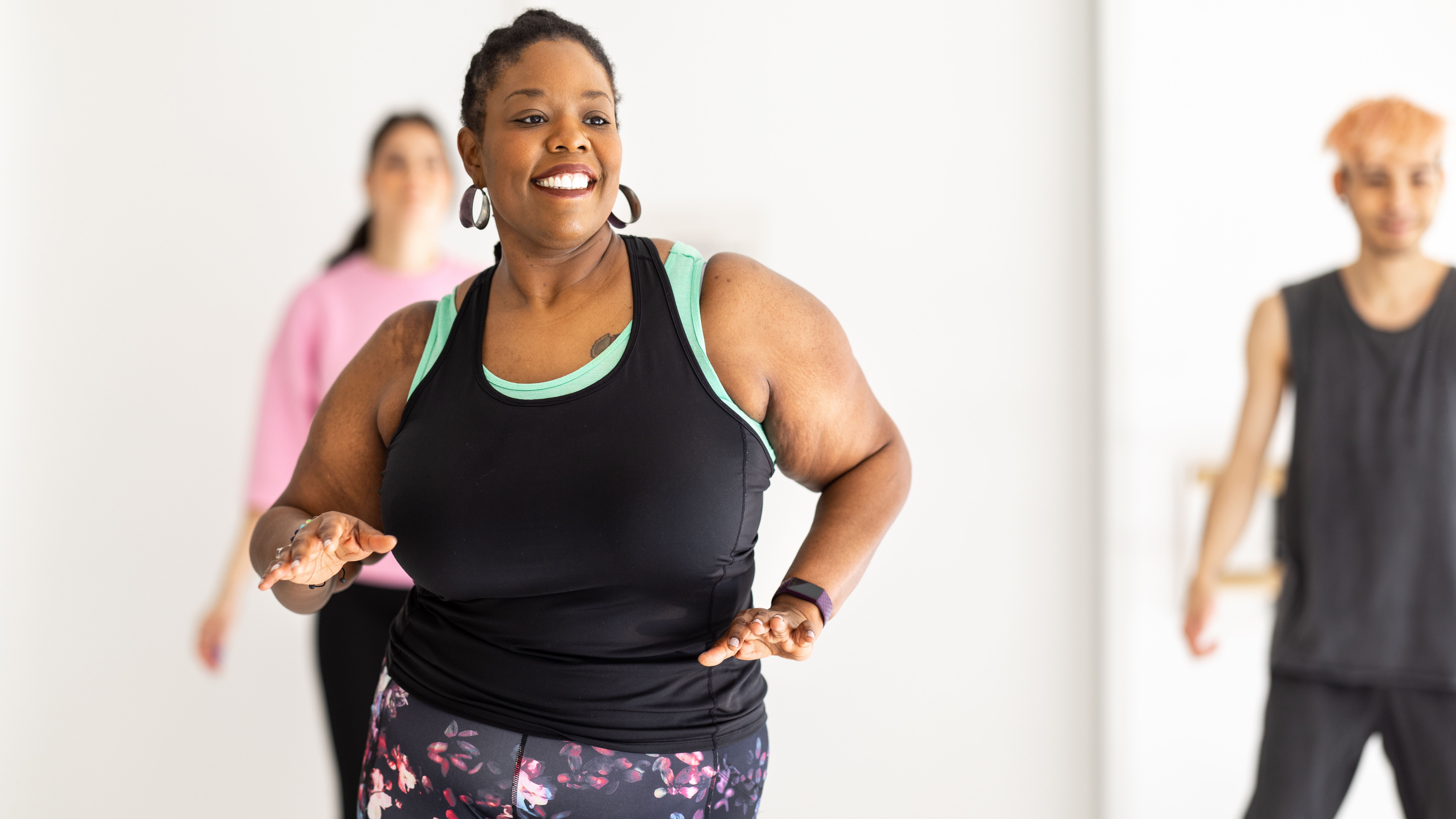 A smiling woman dances in a white studio. There are two people dancing behind her.