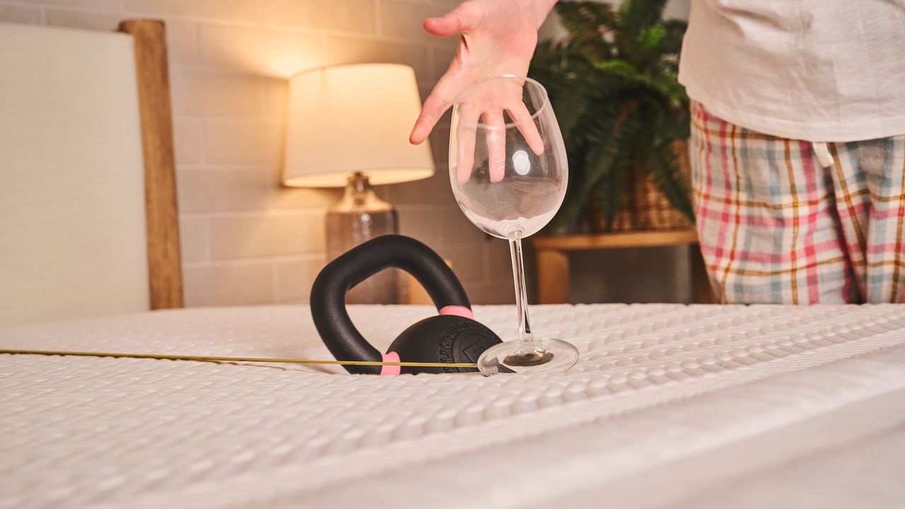 A hand catches a wine glass as it falls down, topped over by a kettlebell that is sinking into the mattress after being dropped onto the Leesa Legend Hybrid Mattress from 4 inches away