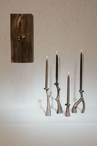 A series of metal candleholder with an abstract, futuristic shape holds black or grey lit candles against a white wall decorated with a brown painting.