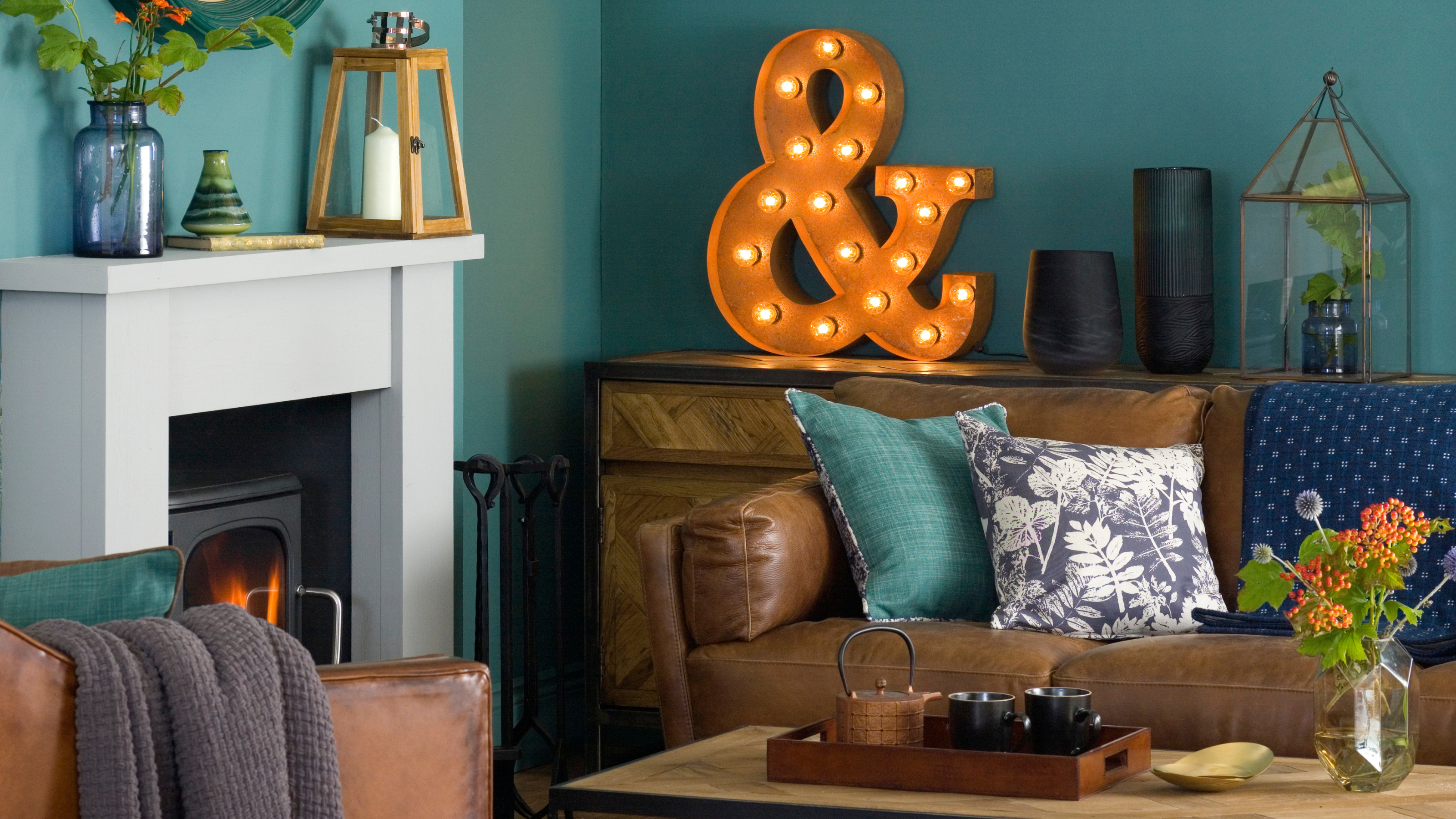 Teal green living room, black woodburner with white mantelpiece, brown leather sofa and amrchair, wood coffee table, large letter light, circular mirror.