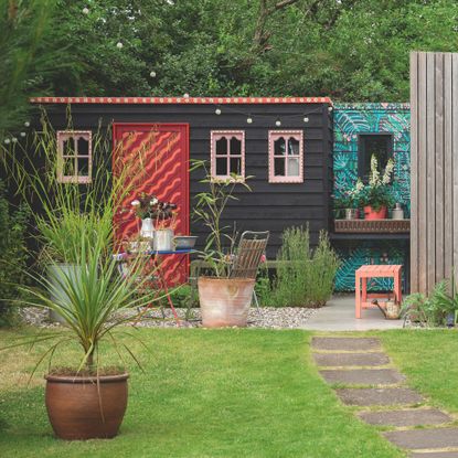 Garden with green lawn area, and a gravel seating area in front of a large black shed with colourful painted door