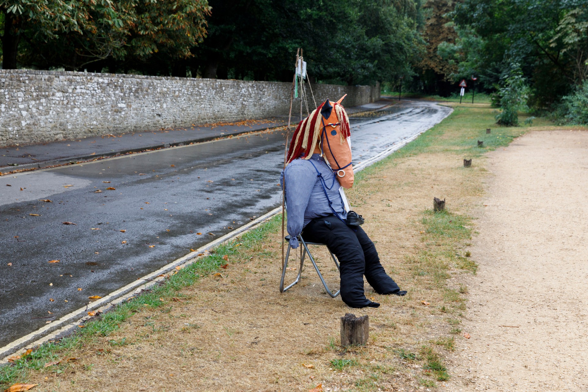 A scarecrow dressed in a blue shirt and black trousers sits on a metal chair at the edge of a quiet, wet road lined with trees. Its head is a handmade fabric horse's head with a rope mane