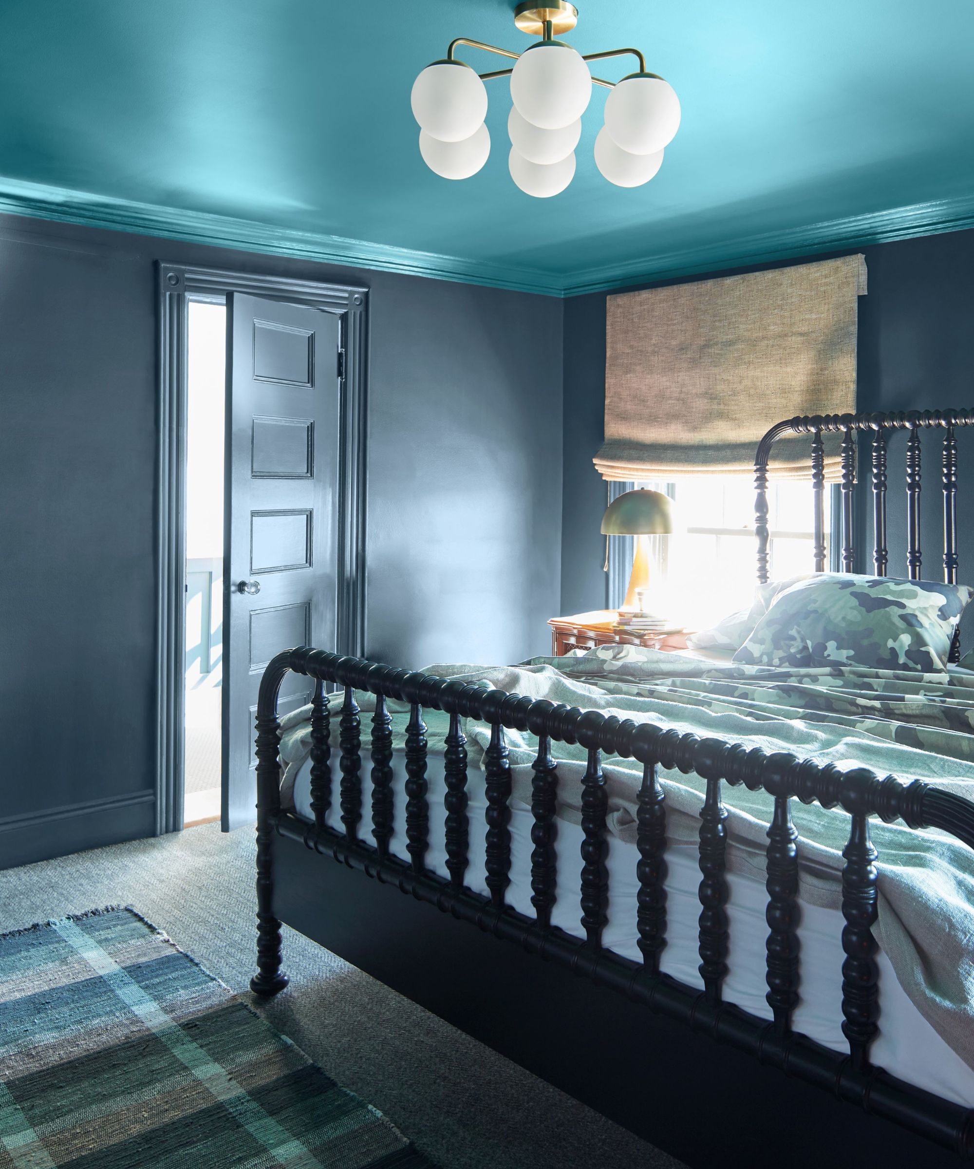 Bedroom with bright blue ceiling, dark blue walls, bobbin frame bed, plaid rug, blue patterned bedding, gold lamp