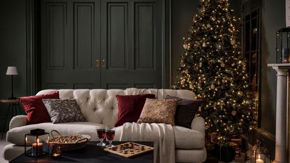 A dark green color drenched living room with a white sofa with red velvet cushions, a well lit tree behind it