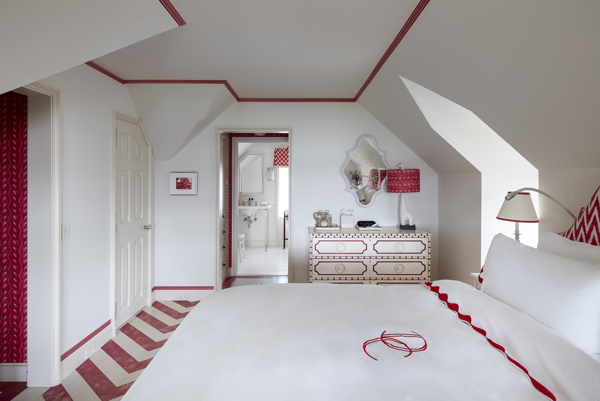 white bedroom with red trims around ceiling and skirting board, white bedding with red scalloped trim, white chest of drawers with red trim detailing, red and white chevron floors, and red wallpaper in hallway