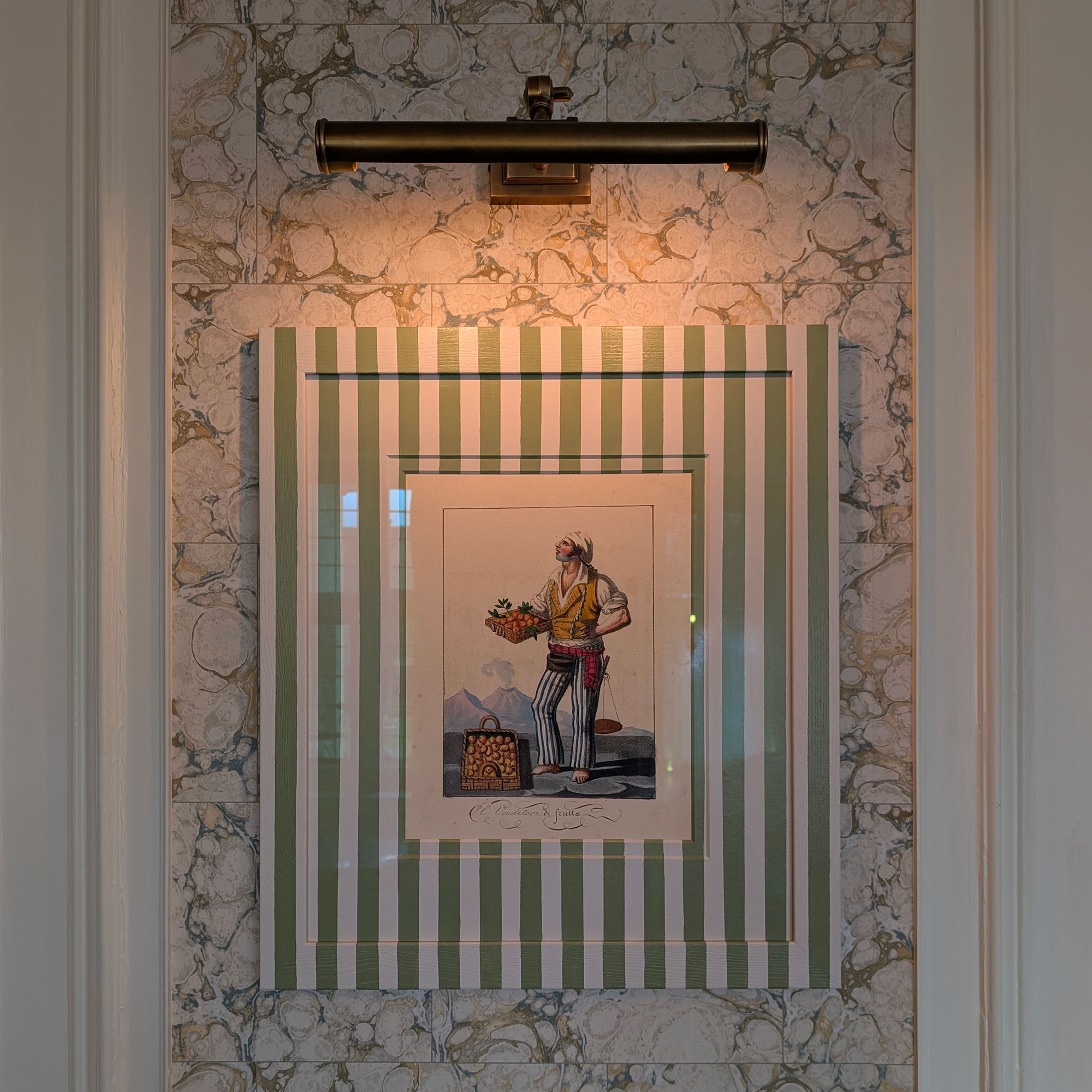 Painting in striped frame on marbled wall underneath brass picture light