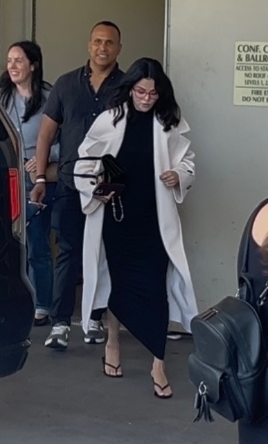 Selena Gomez wearing heeled flip-flops, a black dress, a white coat, and red glasses