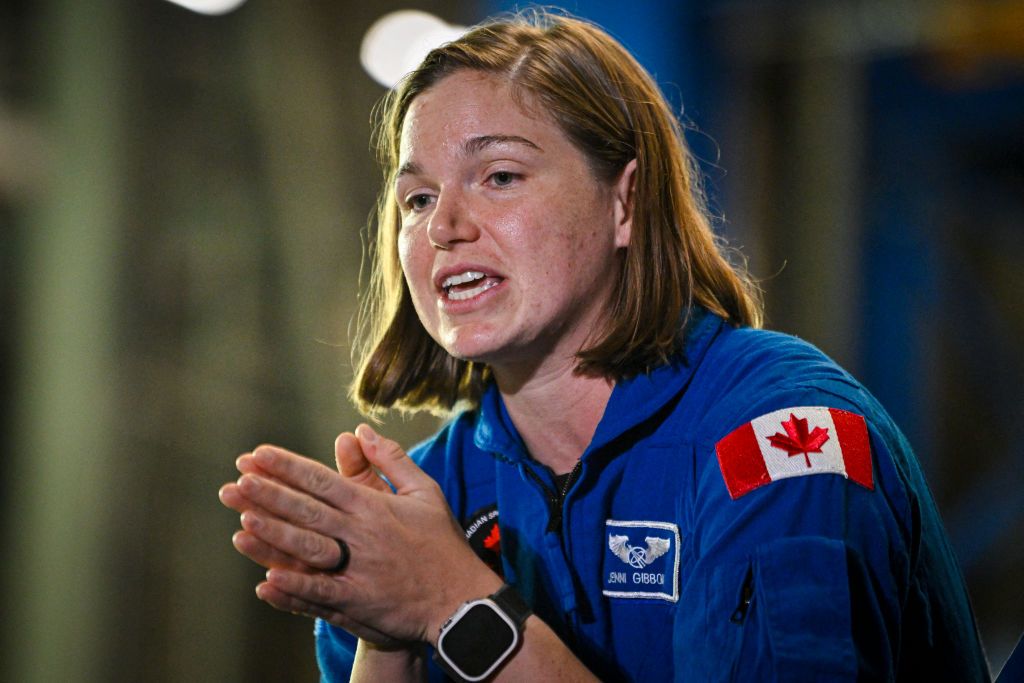 A woman with short brown hair wearing a blue jumpsuit with the red and white Canadian flag on the shoulder looks to the left of the camera.