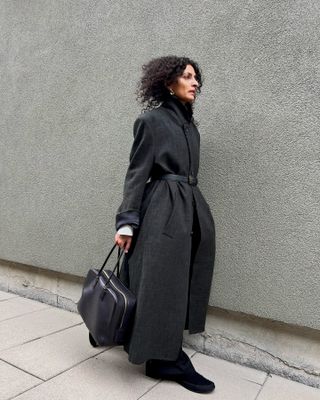 Neelam wears a cinched coat, trousers, black flats, and a black bowler bag.