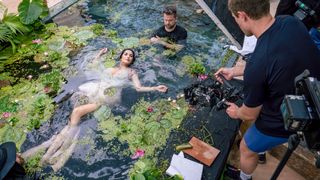 A woman lies in a lake, wearing a silver dress, with a video and photo crew around her, holding cameras and diffusers