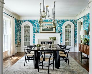 A dining room with turquoise blue and white patterned wallpaper, pendant light with drooping cables, and white arched built-in glass cabinets