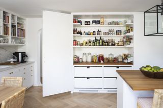 white kitchen with large custom larder with visible shelving and drawers underneath, large doors to each side, both open, kitchen island and cabinets, herringbone floor