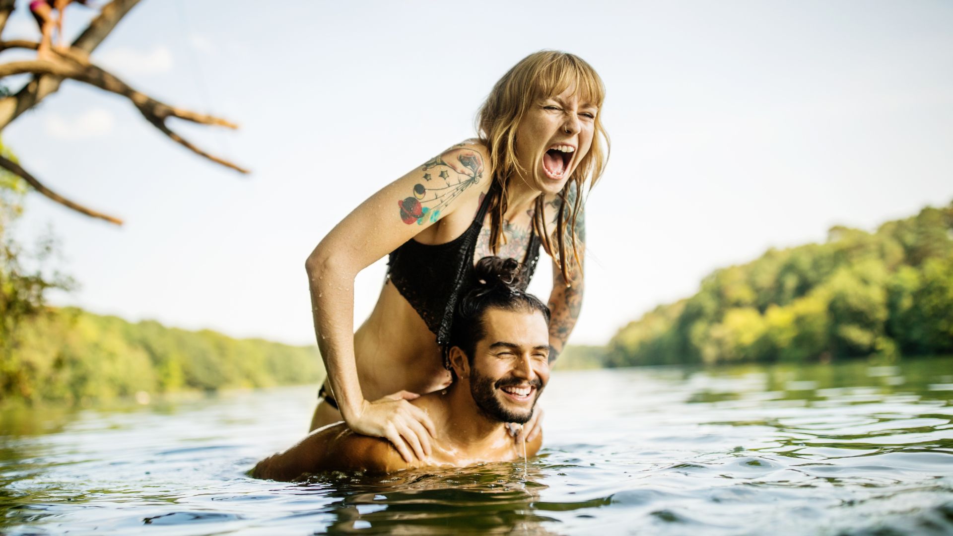 a woman lifts herself up on a man's shoulders as they're both swimming in a river