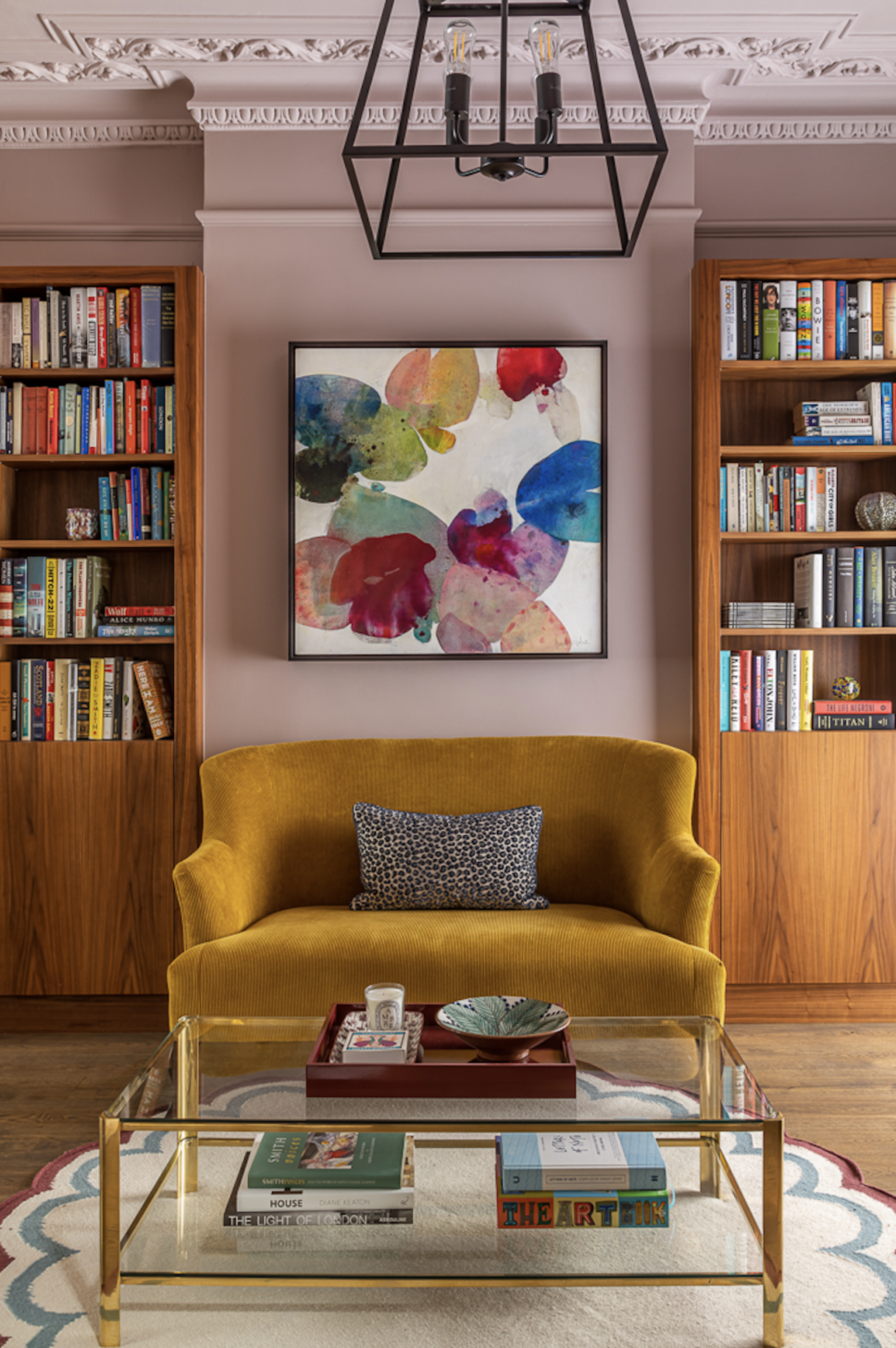 living room with pink walls, yellow velvet sofa, glass coffee table, round rug, colorful artwork and bookcase on either side