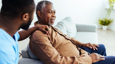 Social worker in foreground rests arm on shoulder of senior man who is sitting on a couch