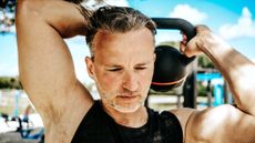 Man in a black vest exercising outside, holding a kettlebell behind his head