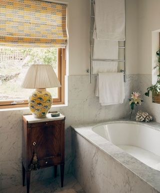 traditional marble bathroom with large window and a small side table with a yellow lamp