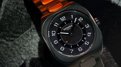 A close-up of the face for a Hermes Paris watch. The colour is dark grey with a dark graphite face, with contains a black circle. The face has white 1-12 numerals with a white hour arm, a black minute arm and a black second arm with a reflective orange triangle. The Hermes Paris logo is in the upper centre of the face with white hashes in the black ring. 