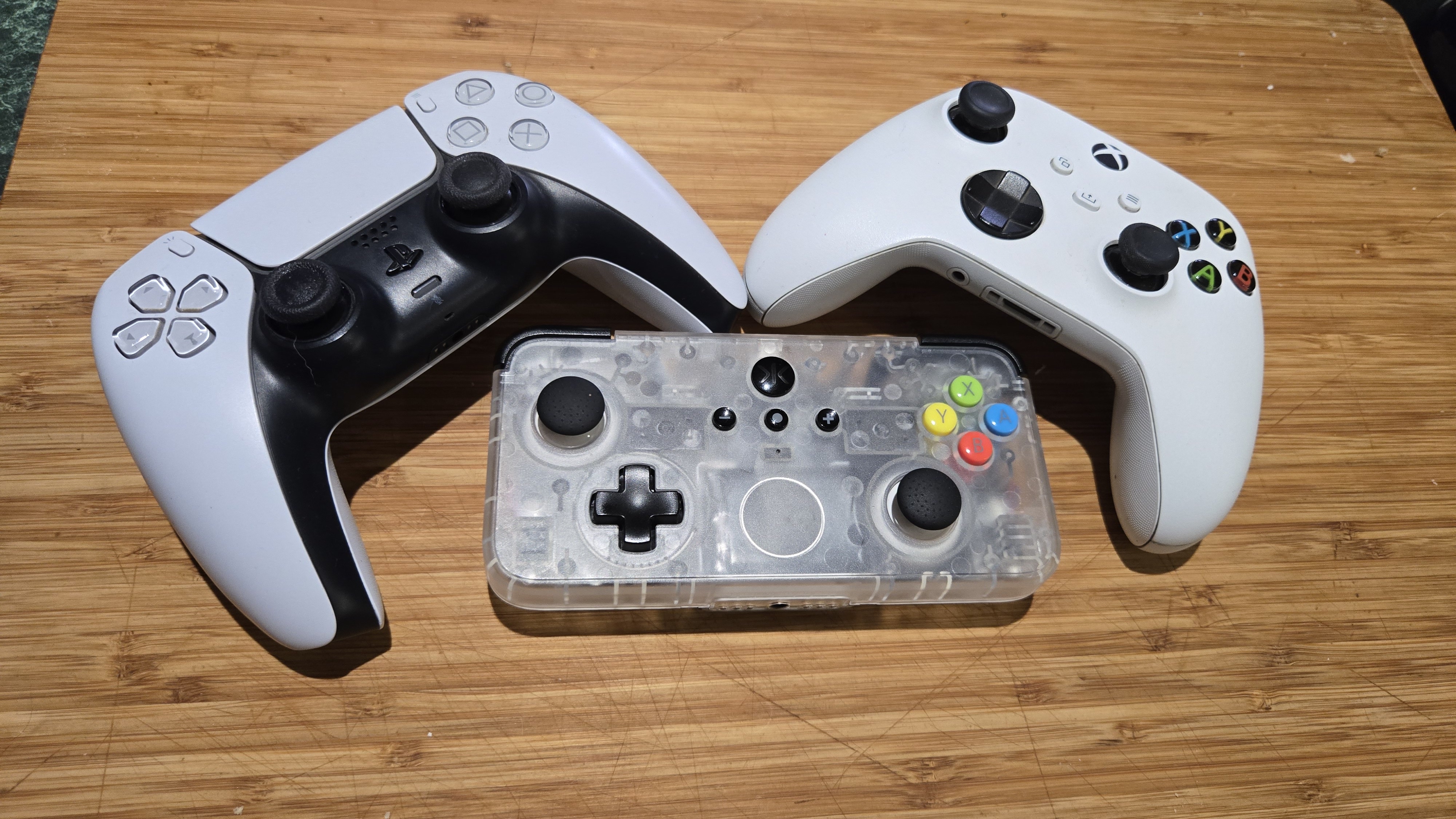 A Classic Clear edition of the CRKD NEO S rectangular controller sits on a wooden countertop next to a Dualsense PS5 controller, and an Xbox controller. The CRKD controller is about half the size of the other two gamepads. The body of the CRKD controller is made from a cloudy clear plastic that allows the viewer to see the internals.