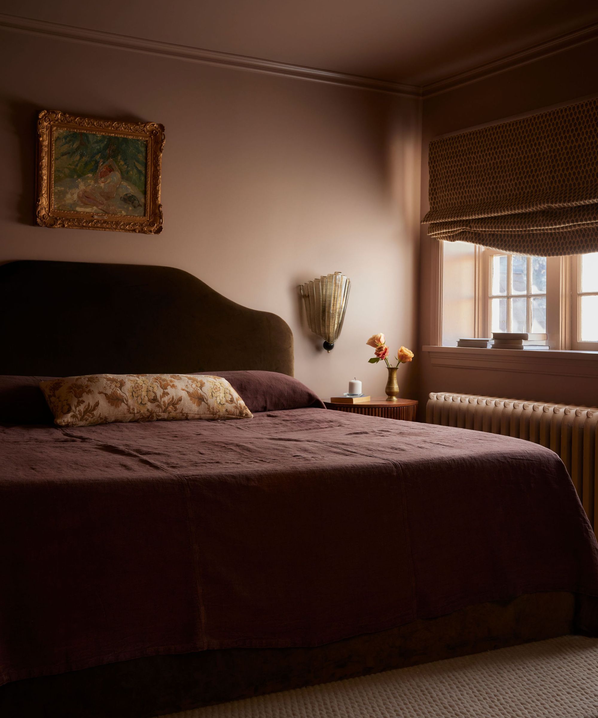 Moody pink and purple bedroom. Large bed with green velvet headboard, purple bedding and floral throw pillow, artwork above bed. Bedside table with vase of flowers, wall light above