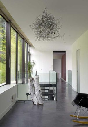 Interior room, white walls and ceiling, grey gloss floor, grey bucket rocking chair, black framed widows, doorway, top of black stairwell with glass barrier, white log sculpture, wire design ceiling light, potted plant, outside view of trees