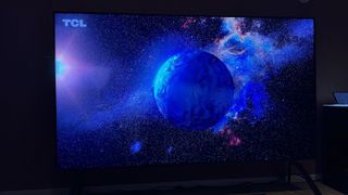 TCL 115QM89 Mini LED TV photographed in a hotel room. On the screen is the image of the Earth and a bright constellation.