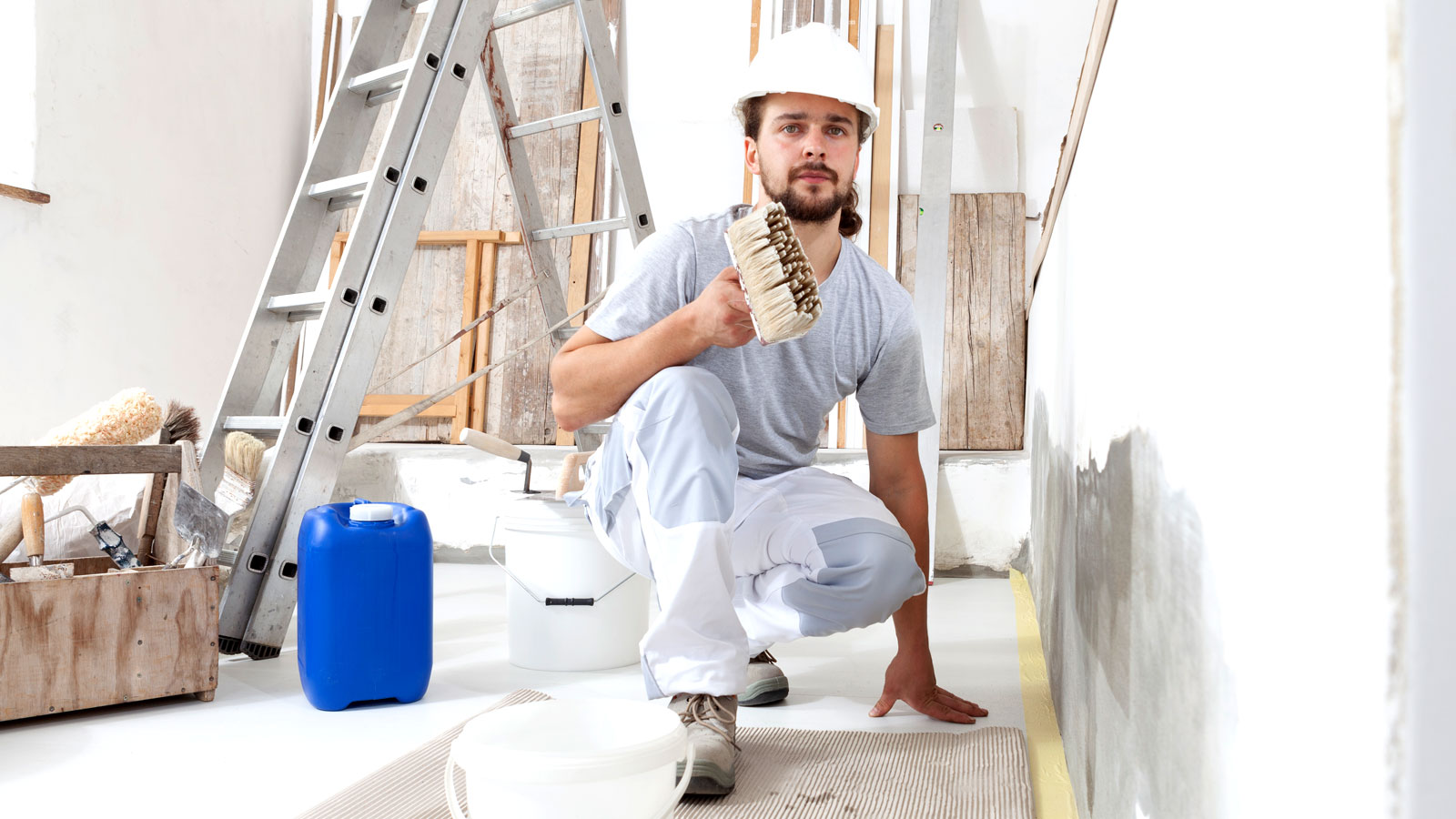 Man in hard hat and overalls with wide paint brush next to unpainted wall with ladder and tools in background