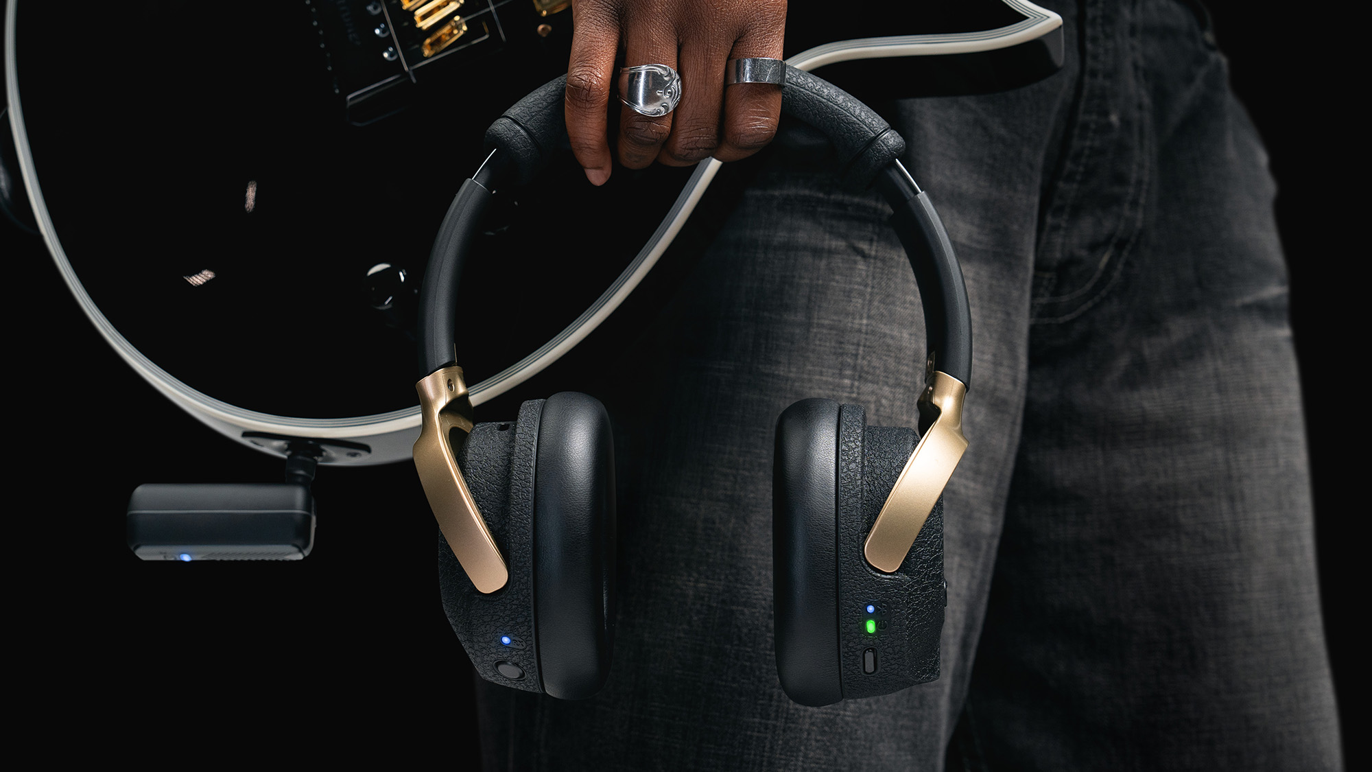 A photo showing a hand holding the Positive Grid NEO headphones next to a black guitar with the NEO dongle inserted into the output jack