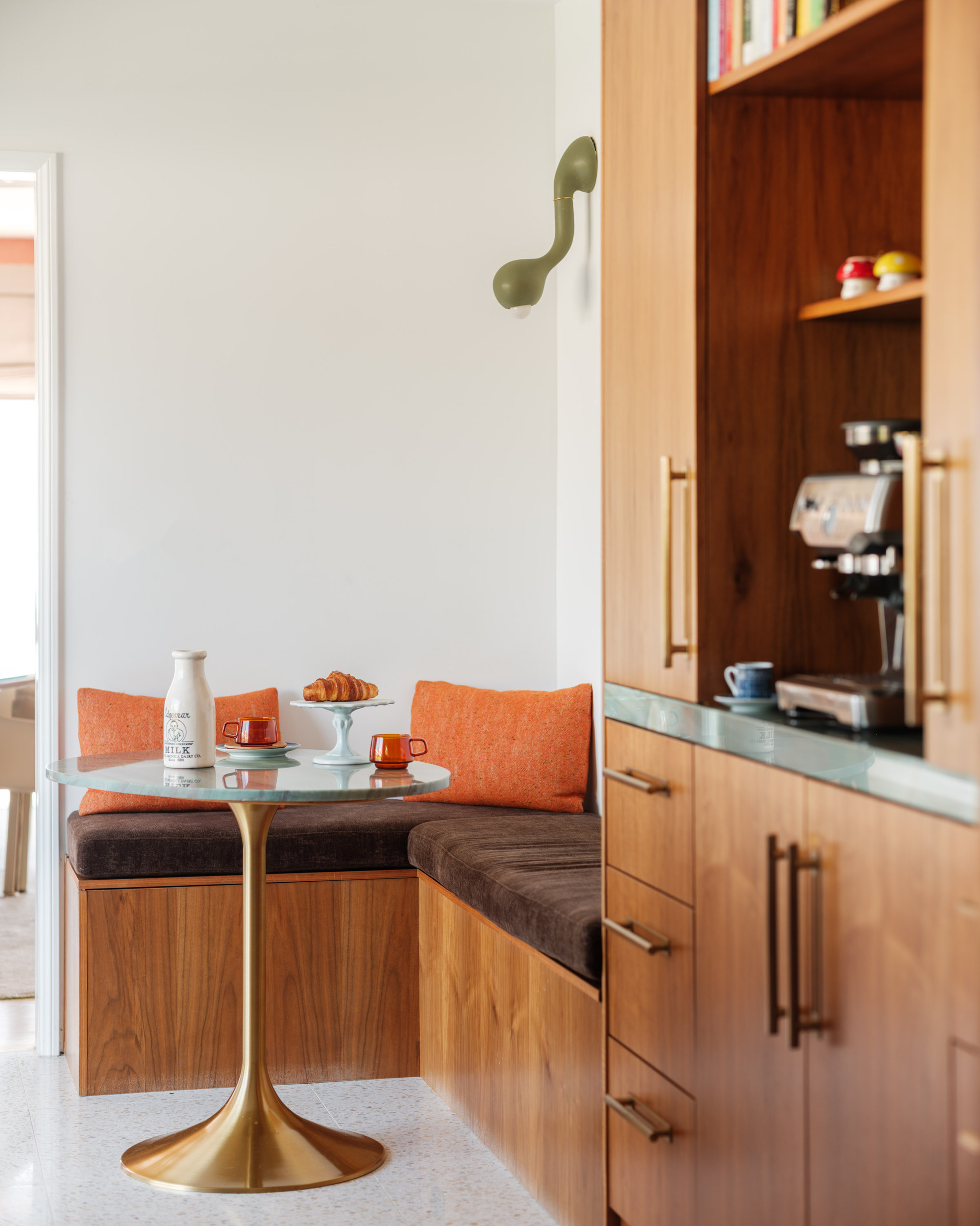 Image of a bright coffee nook/breakfast nook with a warm wooden bench seat that has brown and orange cushions and a small round teal marble table with a golden base.
