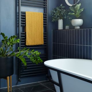 A close-up shot of a black metal heated towel rail with mustard yellow chevron detail towel, white bath with black metal detail, shelf with indoor houseplants in white vases and large black planter with wooden legs
