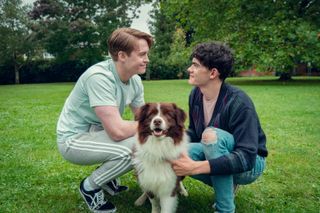 Kit Connor as Nick and Joe Locke as Charlie, smiling at each other as they kneel and pet a dog in a park, in 'Heartstopper' season 3.