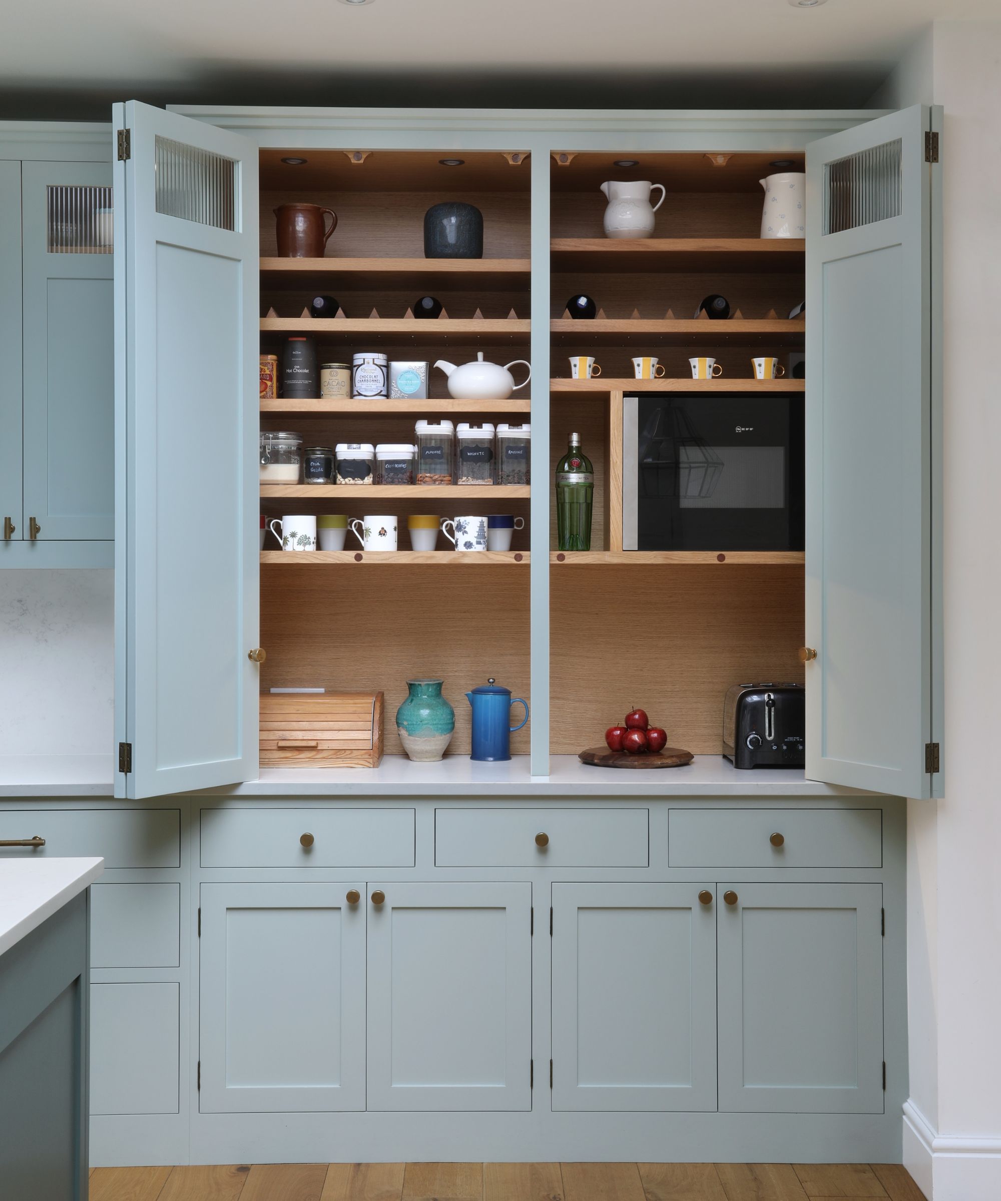 A built-in kitchen pantry with pale blue cabinet doors opened to reveal oak shelves neatly organised with mugs, jars, teapots, and a microwave, combining storage and display.