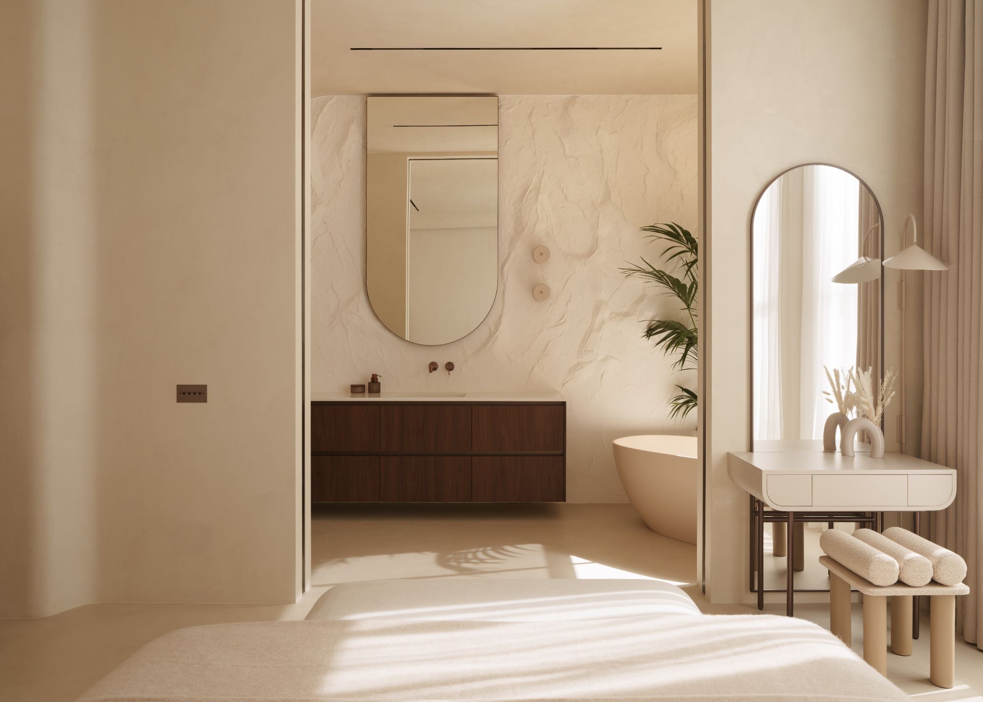 A neutral bedroom with a dressing table and en suite, where the sliding doors are fully open, revealing a dark wood vanity with a large mirror above, a textured cream wall, and the side of a bath
