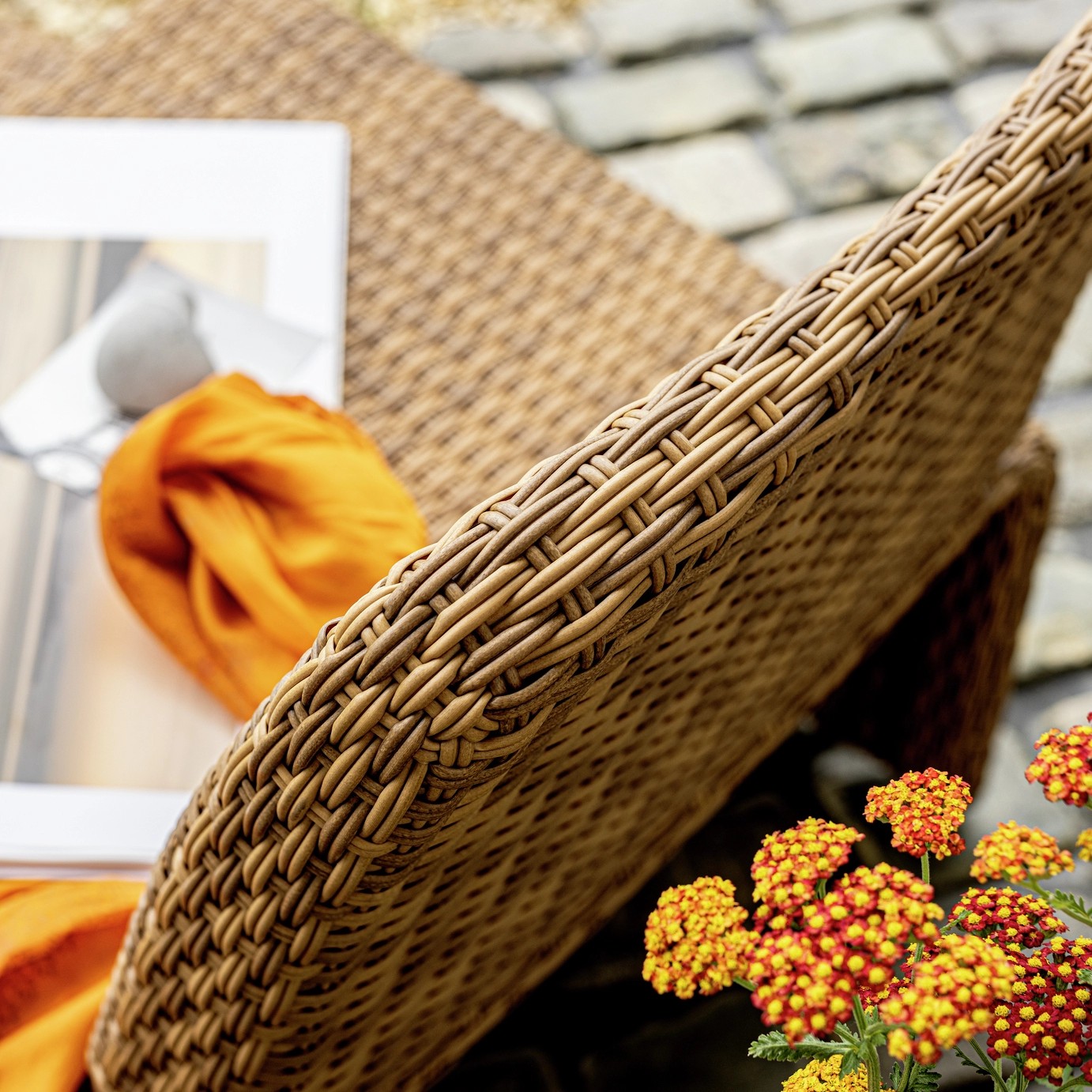 Close-up image of a woven outdoor armchair that shows off the texture of the seat. There is a yellow and red flower by the chair.