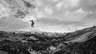 Black-and-white image of a person balancing on a tightrope above a rugged, snow-covered mountain landscape