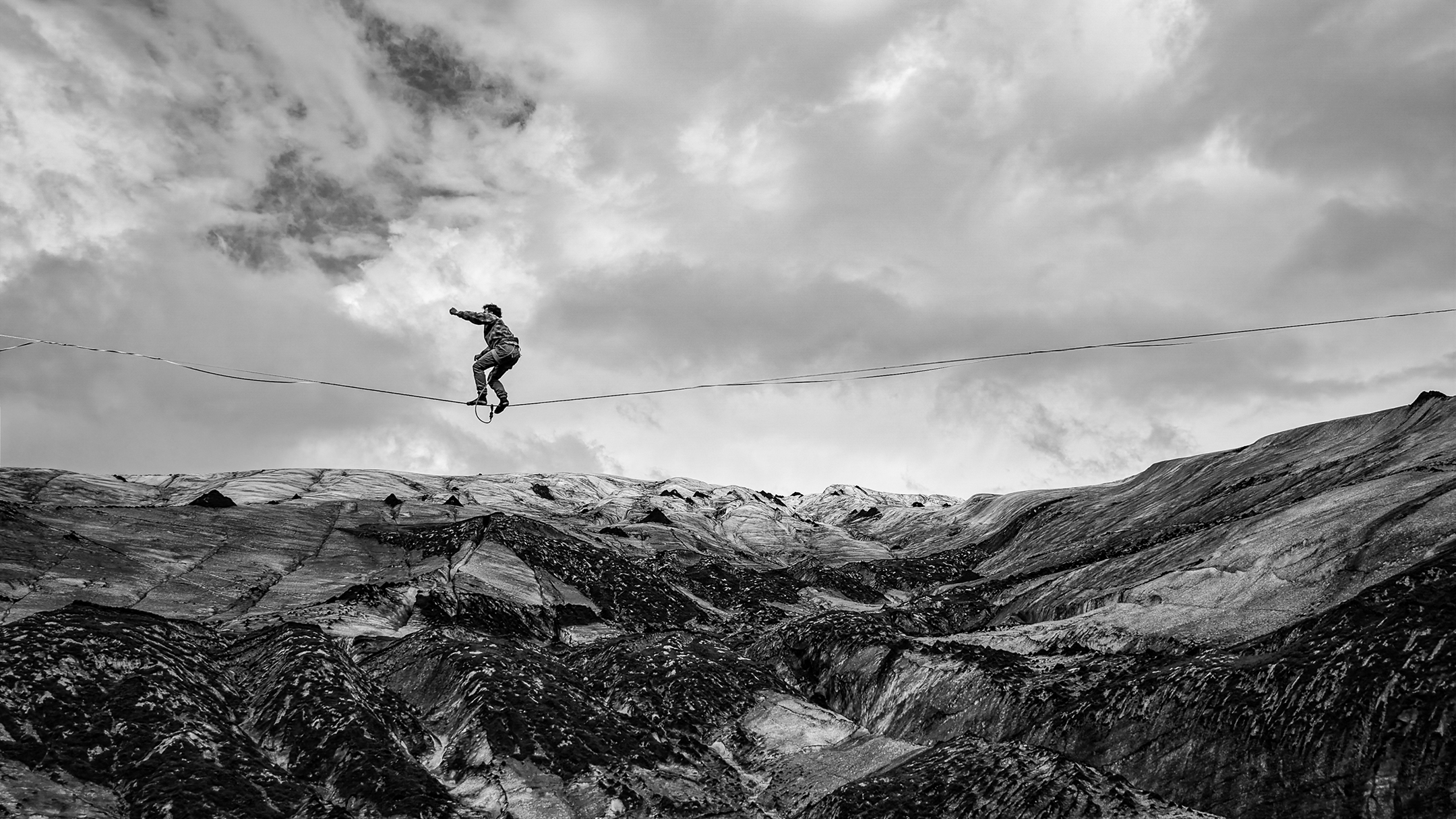 Black-and-white image of a person balancing on a tightrope above a rugged, snow-covered mountain landscape