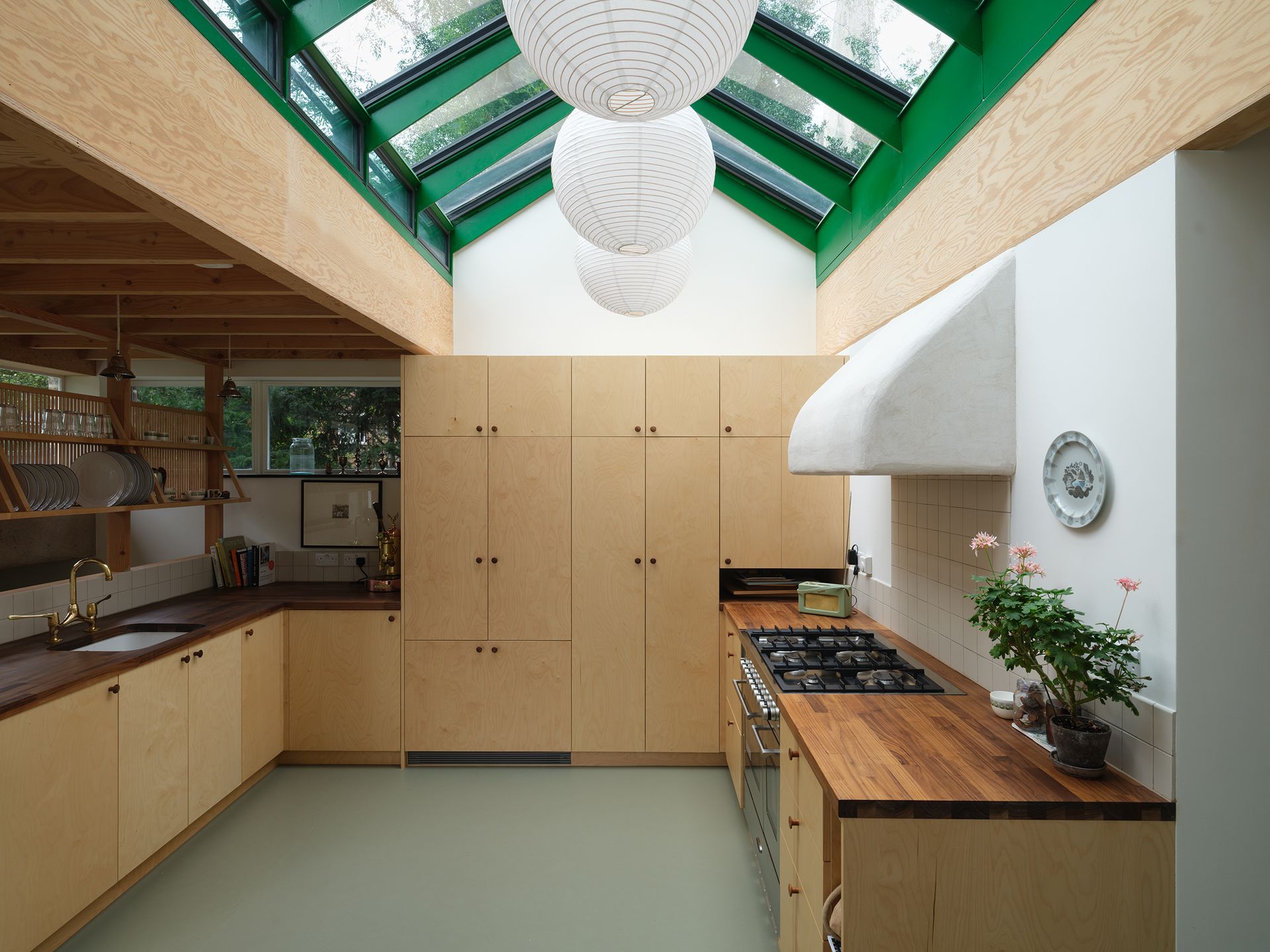 Interior of north London home extension with green skylight, light wood beams, white walls, pale green floor, three large white paper ball ceiling light shades, light wood cupboards and kitchen units, wooden worktops, potted plant, white sink with gold taps, white wall tiles, wooden pots and glasses rack, window, books, gas hob and oven