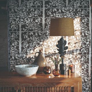 A living room sideboard cabinet with a table lamp and decor objects on top set against a botanical-print wallpaper