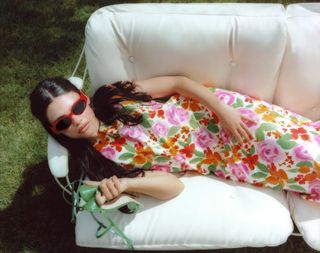 Ariana Greenblatt wearing floral multicolor dress and red sunglasses, laying down.