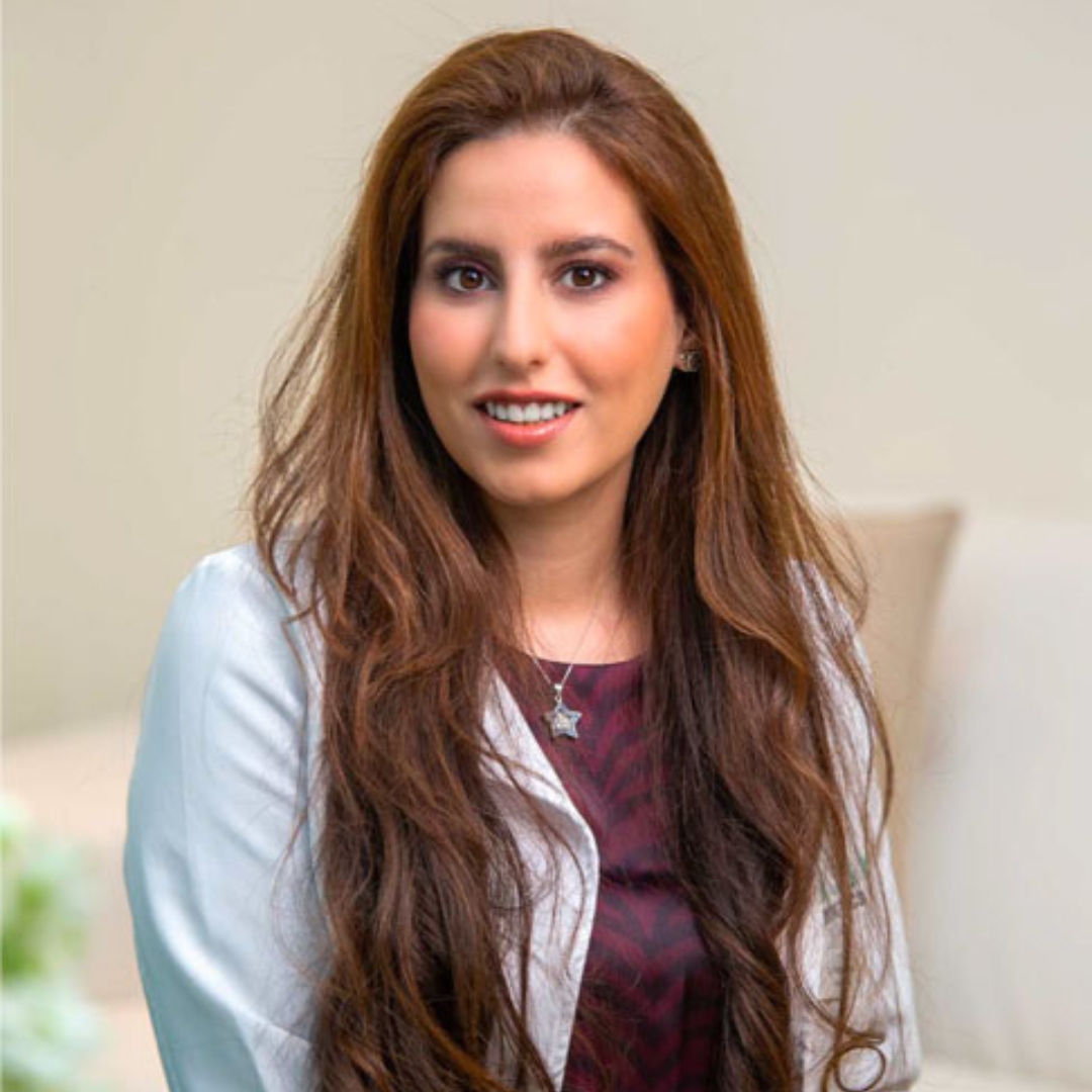 Image of Dr. Asima Nasir smiling with long brunette wavy hair and wearing a burgundy patterned top and a white jacket