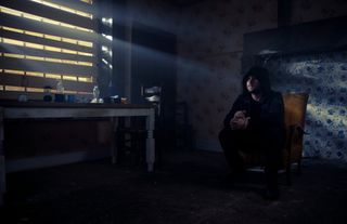 Aaron Dingle in a hood as he sits in a dark abandoned room on a chair.