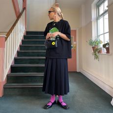 Alfred wears black tee , pleated skirt, Mary Janes, and bright pink socks. 