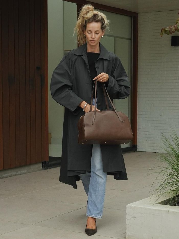 Woman wears trench coat, black top, blue jeans and brown leather bag