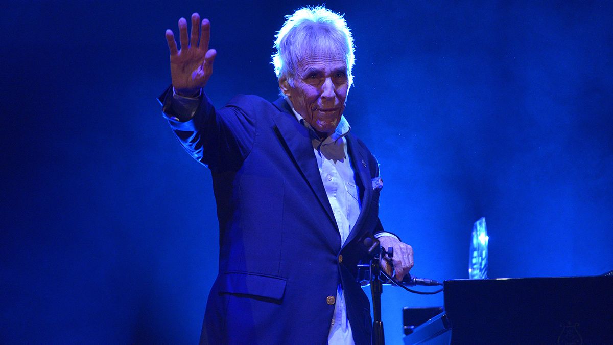 Burt Bacharach stands waving next to a piano