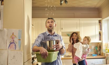 A father tries to catch water leaking from the ceiling with a bucket as his family looks on.