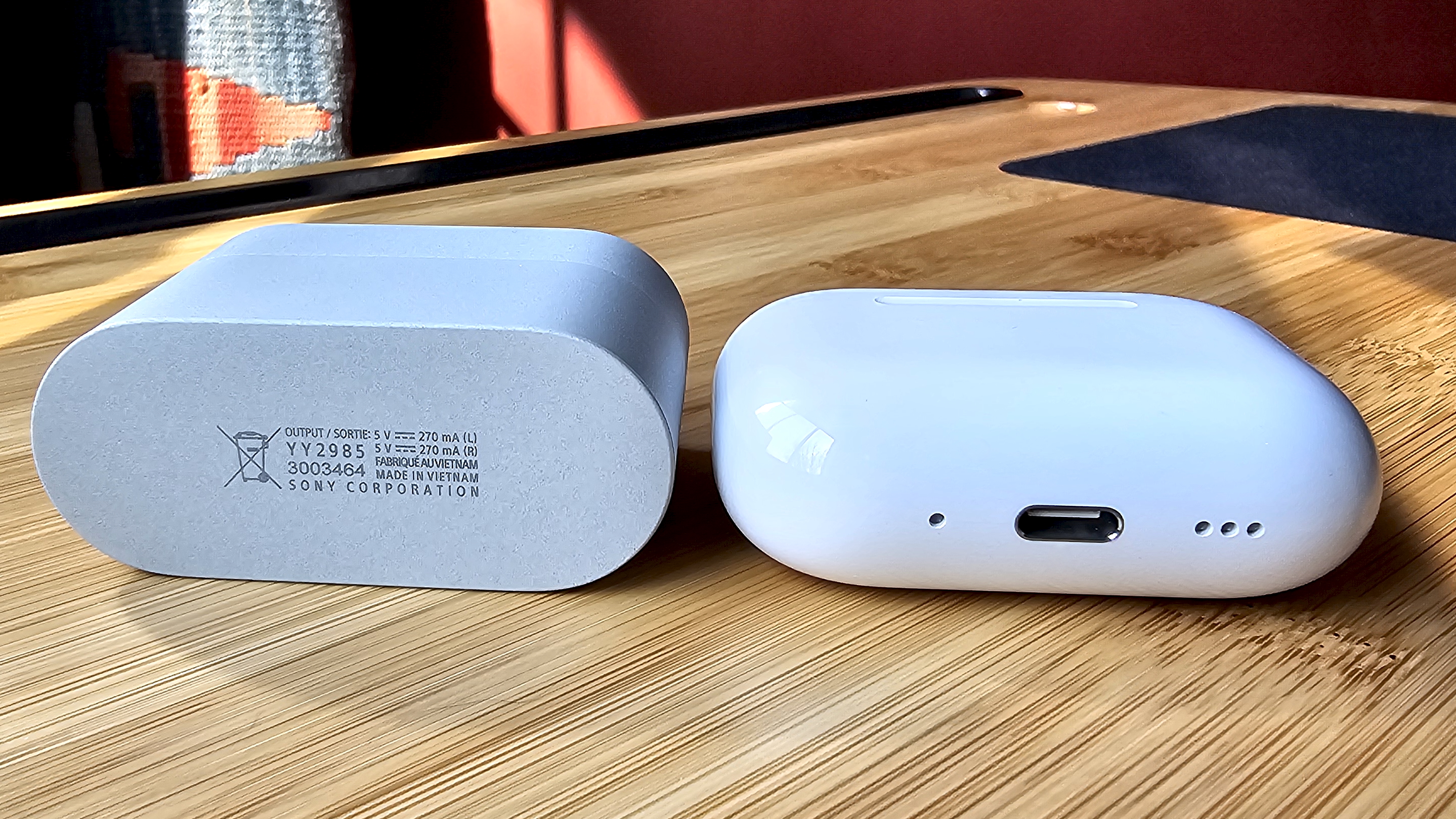 Sony WF-1000XM6 earbuds next to the Apple AirPods Pro 3, on a wooden table, in a versus-style format