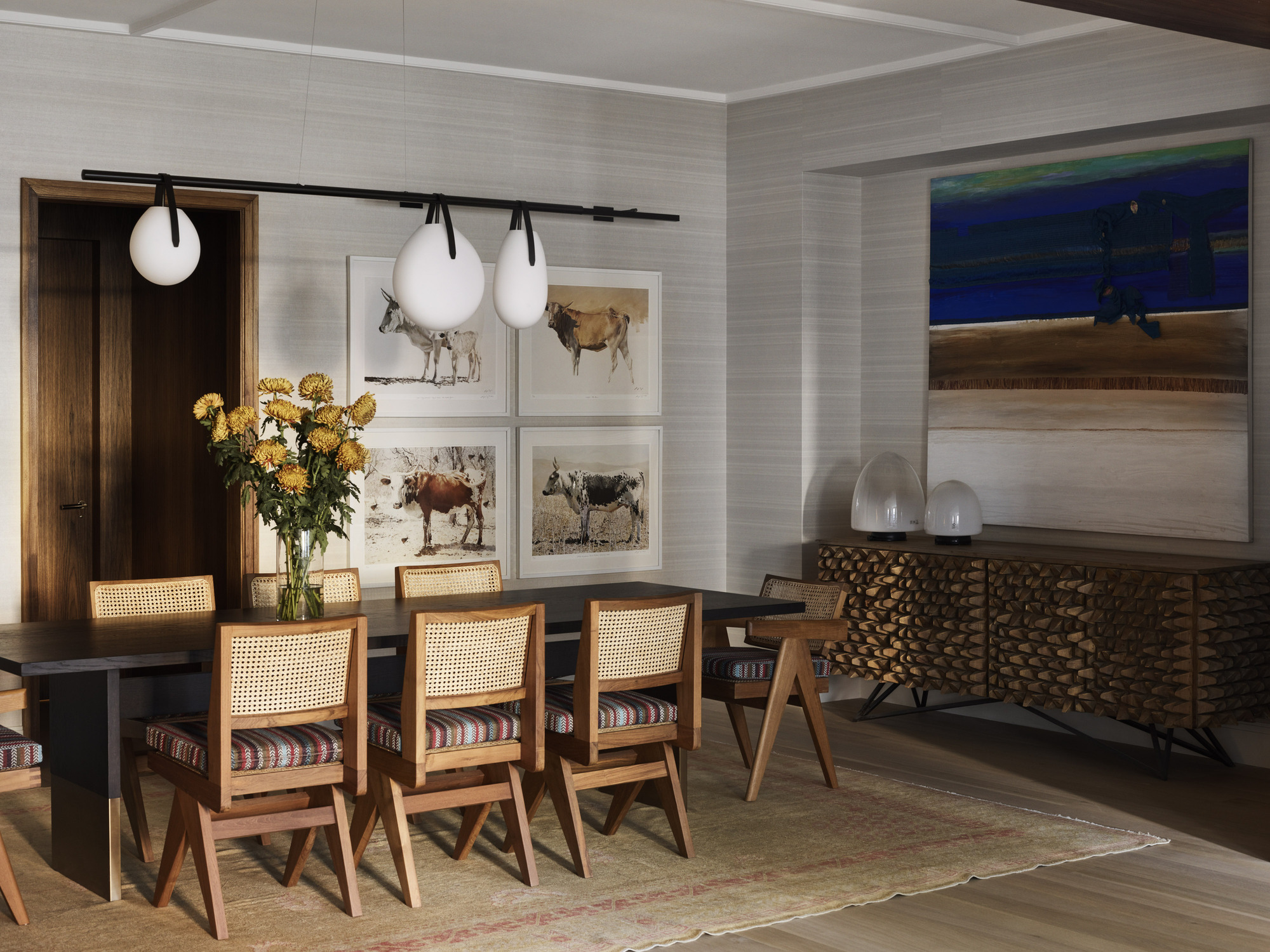 neutral dining room with subtle striped wallpaper, colorful artwork over timber console table, a woven neutral rug under a black timber dining table with timber and woven dining chairs with a long pendant light hung above