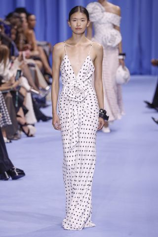 a model walks the runway at Balmain wearing a white polka dot dress with black dots and a floral embellishment in the center